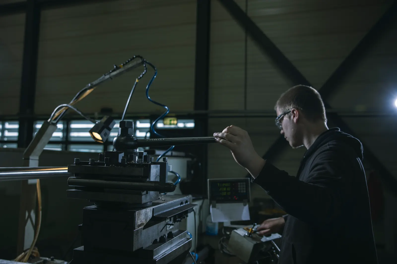 A worker wearing safety glasses operates a conventional lathe with a clamped shaft